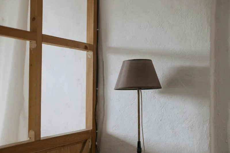 Elegant floor lamp with neutral shade beside a wooden-framed window in a serene room.